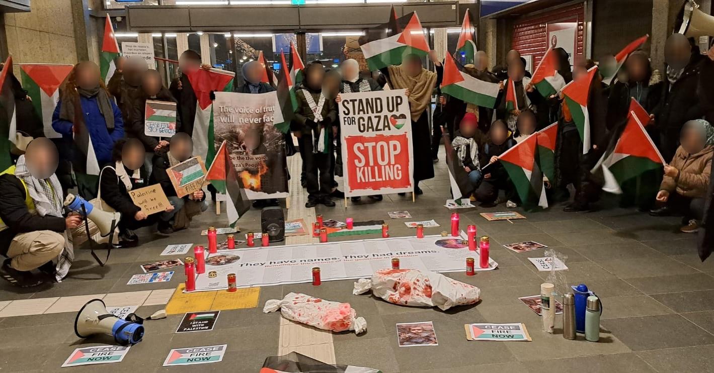 In een verlichte binnenruimte staan en zitten mensen met middelgrote Palestijnse vlaggen en borden. Op de borden staan teksten (in het Engels) waaronder: 'Sta op voor Gaza, stop met moorden', 'De stem van de waarheid zal nooit vervagen', 'Palestina zal vrij zijn', en 'Staakt-het-vuren nu'. Op de grond ligt een groot spandoek met daarop namen van mensen die vermoord zijn, met centraal de tekst (ook in het Engels) 'Ze hebben namen. Ze hadden dromen.' Op de grond staan ook kaarsen, thermosflessen en beelden die lichamen van vermoorde kinderen voorstellen. 