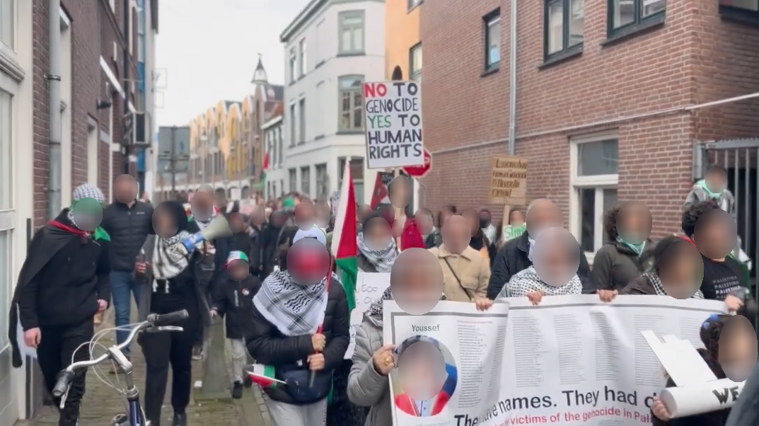 Mensen lopen door een beschutte straat in een binnenstad met spandoeken, borden en Palestijnse vlaggen. Op borden en spandoeken staat onder meer (in het Engels): 'Nee tegen genocide, ja tegen mensenrechten' en 'Zij hebben namen. Zij hadden [dromen]'. 