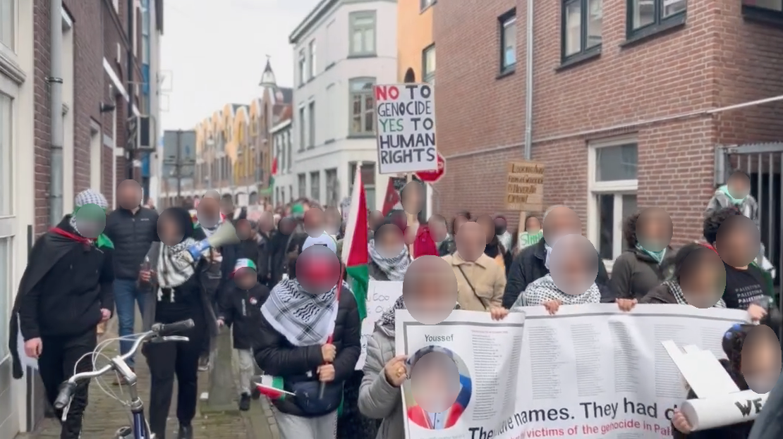 Mensen lopen door een beschutte straat in een binnenstad met spandoeken, borden en Palestijnse vlaggen. Op borden en spandoeken staat onder meer (in het Engels): 'Nee tegen genocide, ja tegen mensenrechten' en 'Zij hebben namen. Zij hadden [dromen]'. 