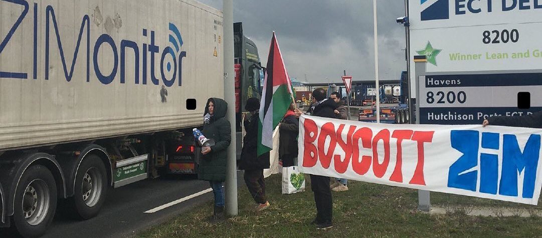 Personen staan in de haven van Rotterdam, bij havens 8200, met Palestijnse vlaggen, flyers, en een spandoek met daarop de rood-blauwe tekst, in het Engels: 'Boycott Zim'. 
