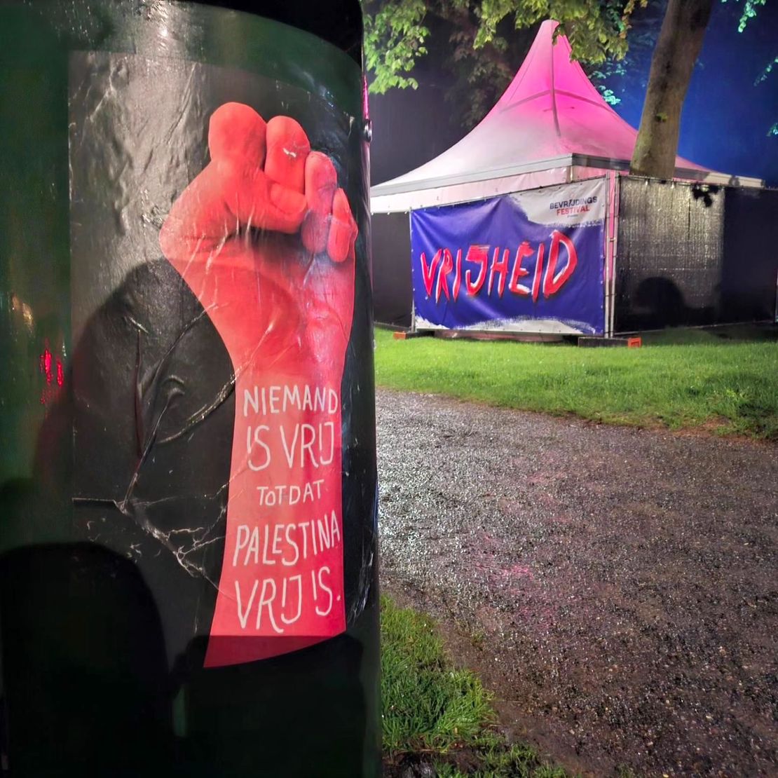 Een nachtscène met Op de voorgrond lantaarnlicht een donkergroene zuil met daarop een poster met een rode vuist en in wit op de pols de tekst: 'Niemand is vrij totdat Palestina vrij is'. Op de achtergrond een roze-verlichtte, witte feesttent achter een hek, met daarop een spandoek met in grote letters het woord 'Vrijheid'.