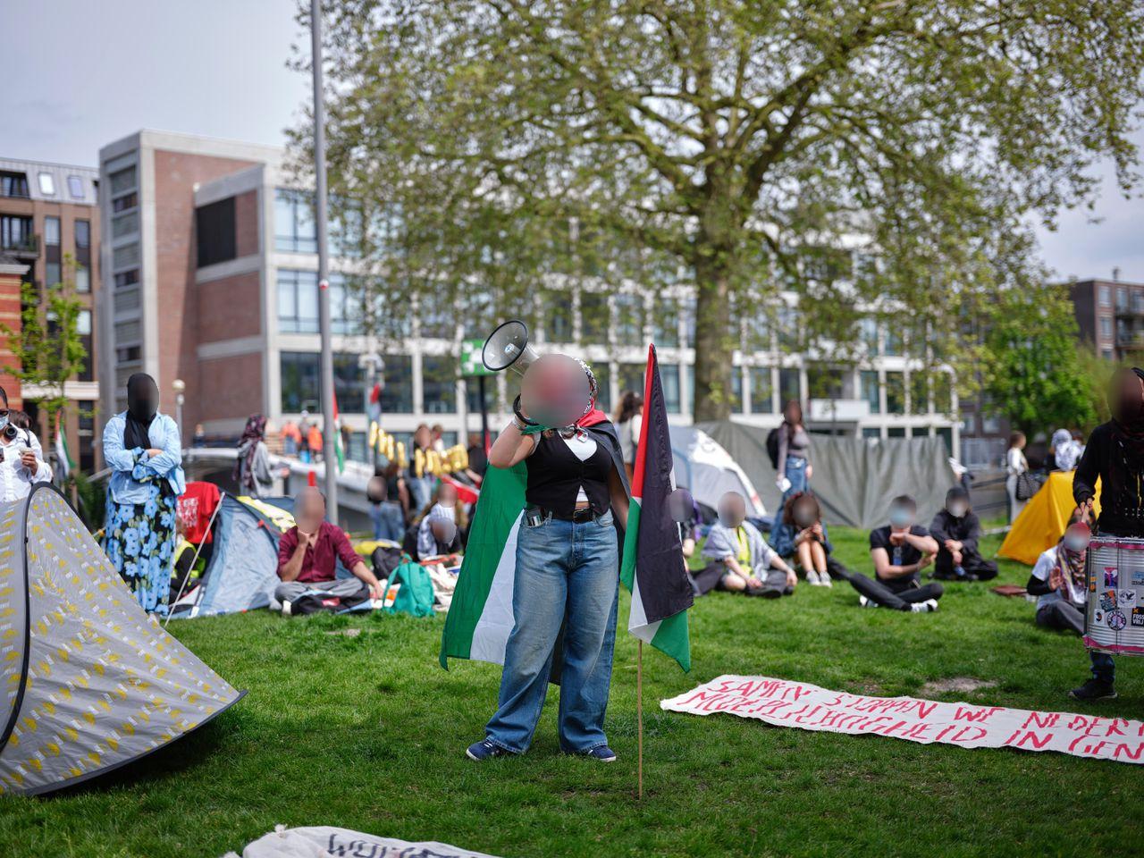 Een persoon staat op een grasveld en spreekt een groep mensen toe. Ze houden een megafoon voor hun gezicht en dragen een keffiyeh en een Palestijnse vlag als cape. Op de achtergrond zitten andere mensen op het gras, sommigen voor tenten. Op een spandoek op de grond voor de spreker staat 'SAMEN STOPPEN WE NEDER[LANDSE] MEDEPLICHTIGHEID IN GEN[OCIDE]'.