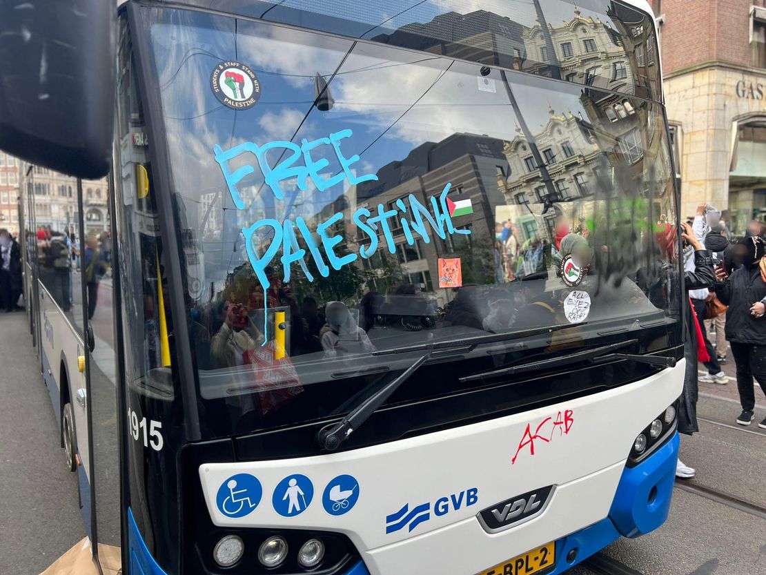 Een wit-blauwe bus van de Amsterdamse vervoerder GVB staat midden op de weg stil, met op de voorruit en voorkant verschillende protestboodschappen. Onder meer staat er in grote blauwe stiftletters 'FREE PALESTINE' ('BEVRIJD PALESTINA'), stickers met een Palestijnse vlag erop, en met rode stift de afkorting 'ACAB' ('Alle politieagenten zijn schurken'). Verschillende mensen staan om de bus heen, waarvan sommigen maskers en keffiyeh dragen.