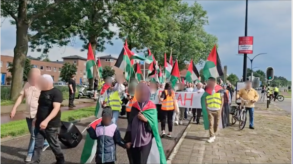 Een stoet met honderden mensen met Palestijnse vlaggen, spandoeken en borden marcheert op een zonnige dag over straat in een open gebied met wat huizen en bomen. 