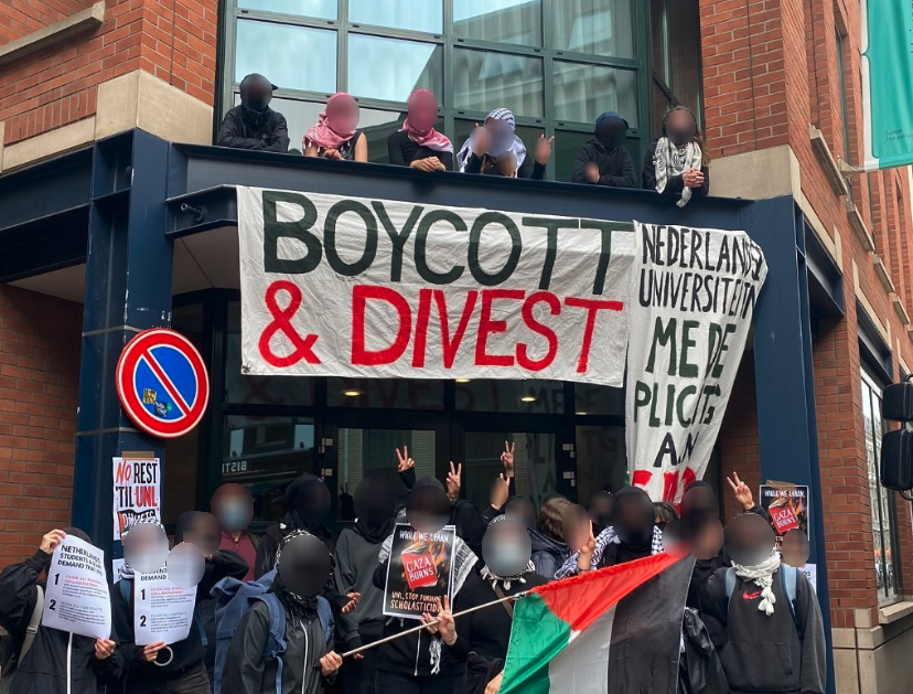 Buiten, bij de voorkant van een bakstenen, stalen en glazen gebouw, staat en zit een twintigtal personen met posters, Palestijnse vlaggen en grote spandoeken met daarop de teksten (in Nederlands en Engels): 'Boycot en desinvesteer', en 'Nederlandse universiteiten medeplichting aan ...' [onleesbaar]. Zes van hen zitten bovenop het portaal van de ingang. Het grootste gedeelte draagt keffiyeh en maakt met hun vingers het 'V'-teken van overwinning. 