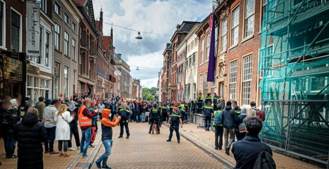 Een grote menigte mensen verzamelt zich midden op straat en langs de trottoirs van een stadsstraat. De straat is geplaveid met lichtbruin-oranje stoeptegels, met aan beide kanten van de straat ​​grote bakstenen gebouwen met meerdere verdiepingen. Eén gebouw aan de rechterkant van de straat is deels bedekt met steigers en een blauwgroen vangnet. Een aantal mensen draagt borden en spandoeken. Vanaf een bovenverdieping van een gebouw aan de rechterzijde van de straat hangt een lang, gedrapeerd spandoek aan de gevel. Er is heel veel politie aanwezig, die zich verzamelt rond de ingang van het gebouw met het spandoek.