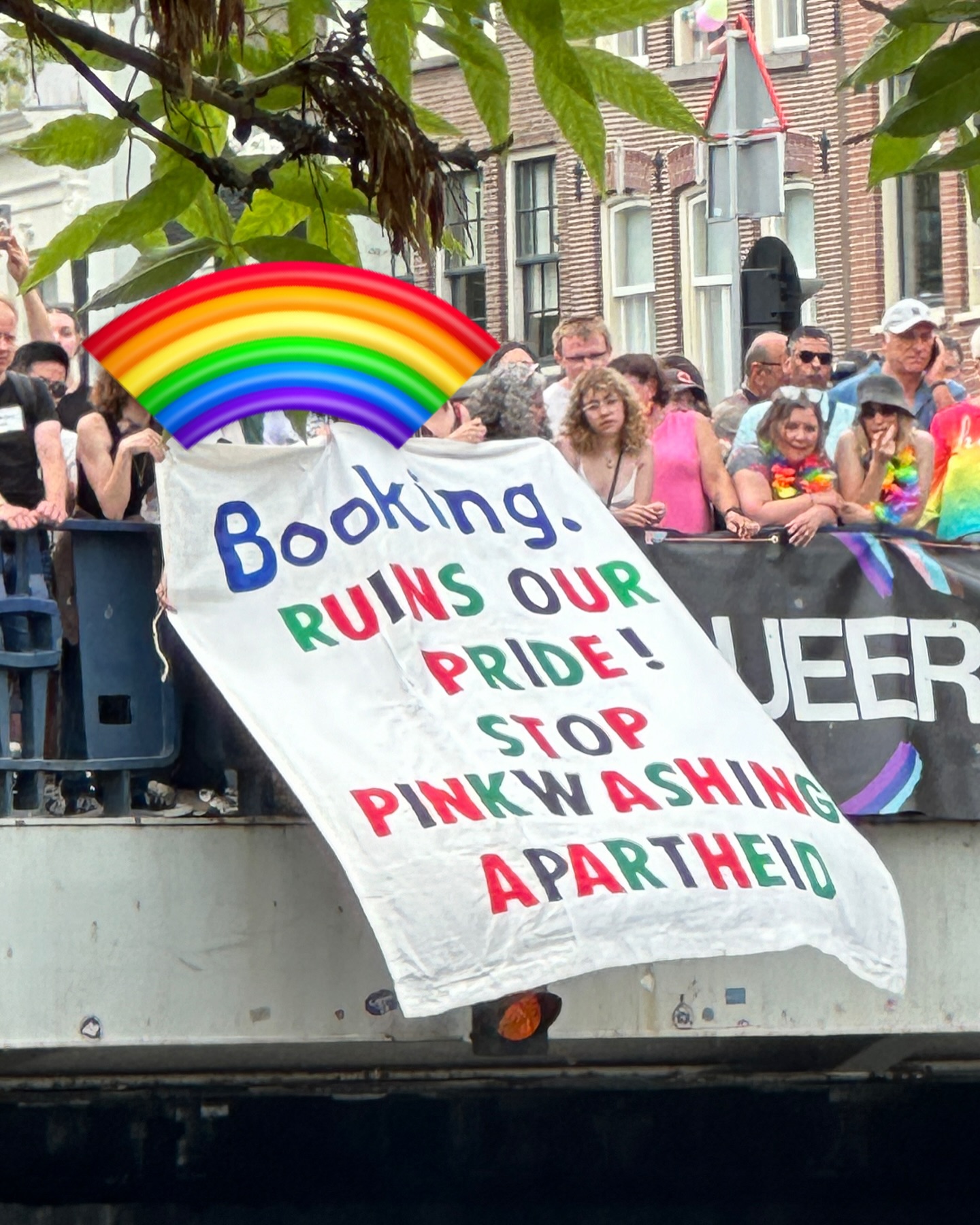 Op een brug staat een massa kleurrijk uitgedoste mensen. In het midden van de brug wordt een spandoek naar beneden uitgeworpen met daarop de tekst, in rode, groene, zwarte en blauwe letters (in het Engels): 'Booking verpest onze Trots[parade]! Stop met het rozewassen van apartheid'.