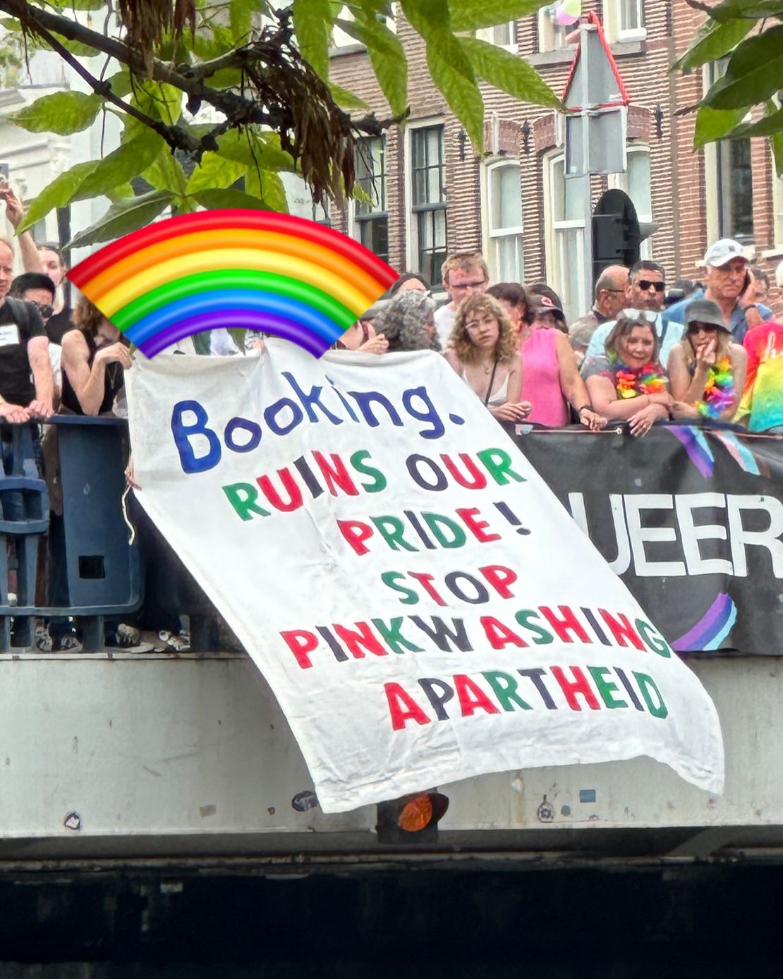 Op een brug staat een massa kleurrijk uitgedoste mensen. In het midden van de brug wordt een spandoek naar beneden uitgeworpen met daarop de tekst, in rode, groene, zwarte en blauwe letters (in het Engels): 'Booking verpest onze Trots[parade]! Stop met het rozewassen van apartheid'.
