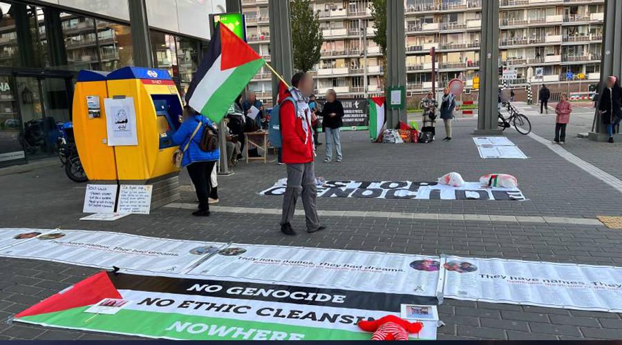 Een groep demonstranten met spandoeken die op de grond liggen, en Palestijnse vlaggen. Er is ook een NS kaartautomaat. 
