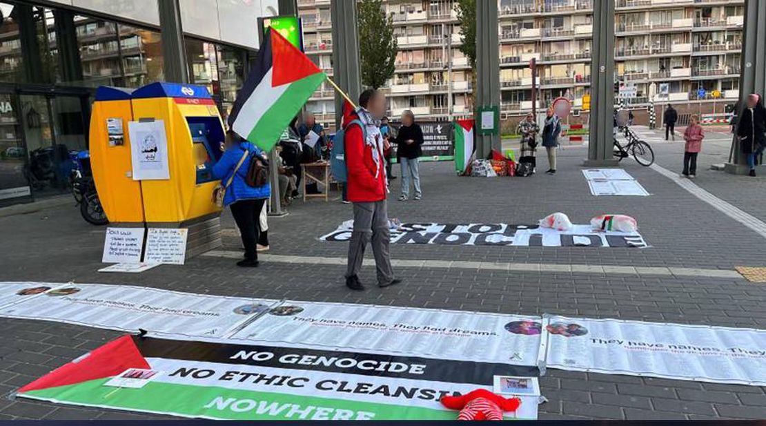 Een groep demonstranten met spandoeken die op de grond liggen, en Palestijnse vlaggen. Er is ook een NS kaartautomaat. 